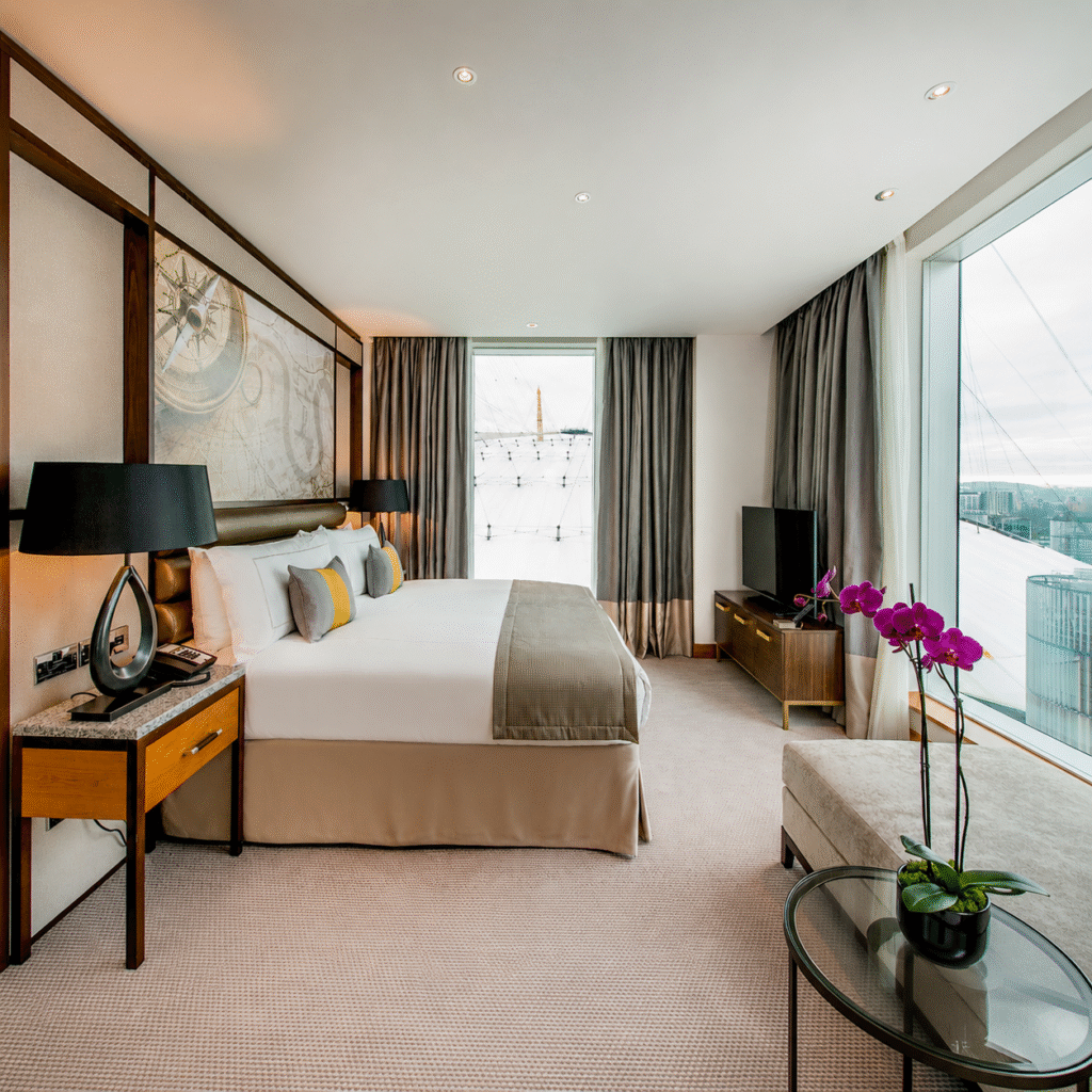King bed in a modern hotel room with lamps, artwork, and a large window overlooking the city; a purple orchid sits on a glass table in the foreground.