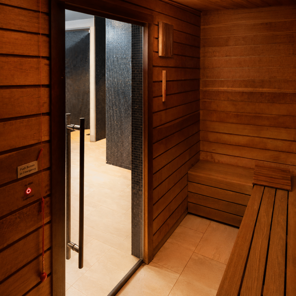 Wood-paneled sauna interior with a slatted bench and open glass-door leading to a tiled shower area; red emergency alarm cord and button on the wall.