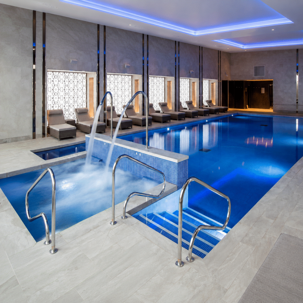 Luxurious indoor spa with a long blue-lit pool, row of lounge chairs along the wall, and a raised hot tub with metal railings.