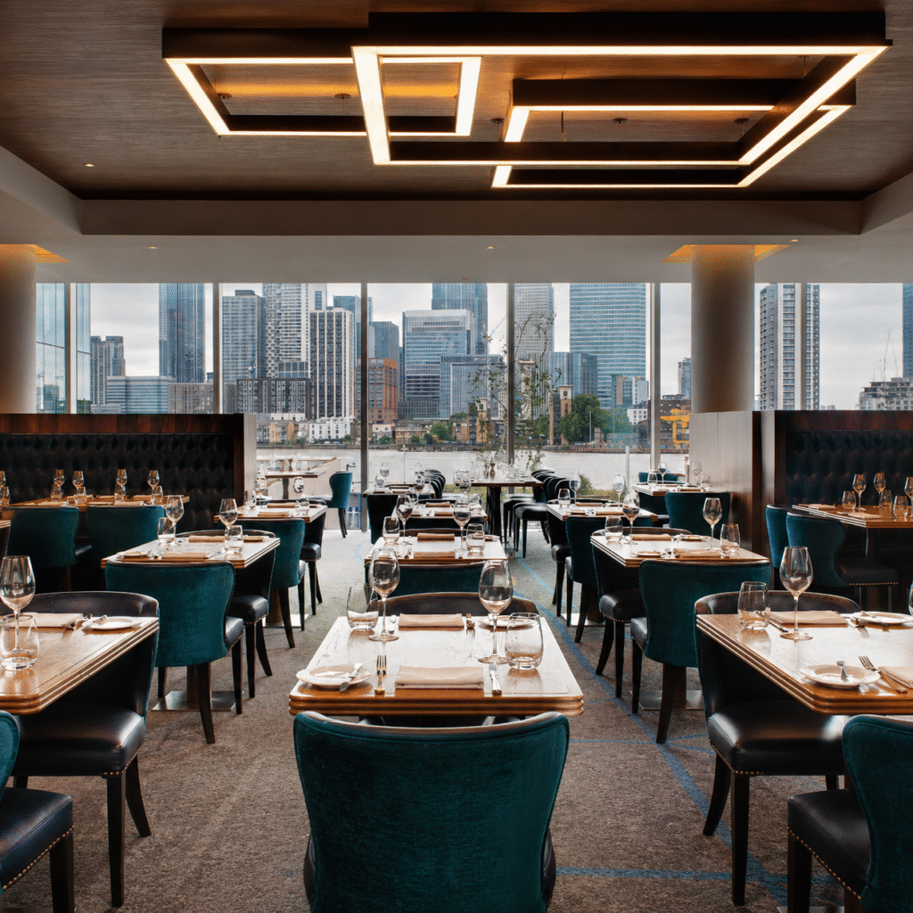 Upscale restaurant dining area with teal upholstered chairs, neatly set tables, and large windows overlooking a city skyline and river.