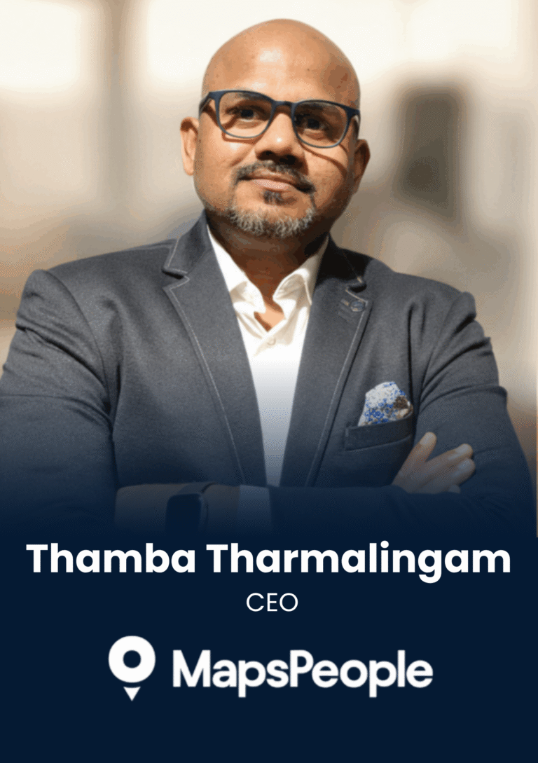 Portrait of Thamba Tharmalingam, CEO of MapsPeople, wearing a navy blazer and glasses, facing camera with arms crossed