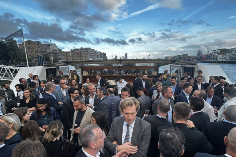 Crowded boat deck with guests in formal attire mingling and holding drinks, city buildings in the background.
