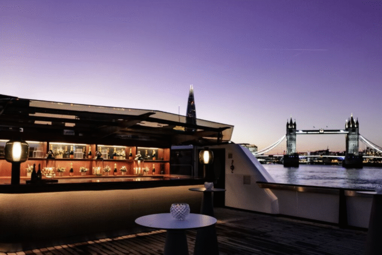 Rooftop bar at dusk with warm lights, seating along the bar and Tower Bridge across the Thames in the distance under a purple sky.