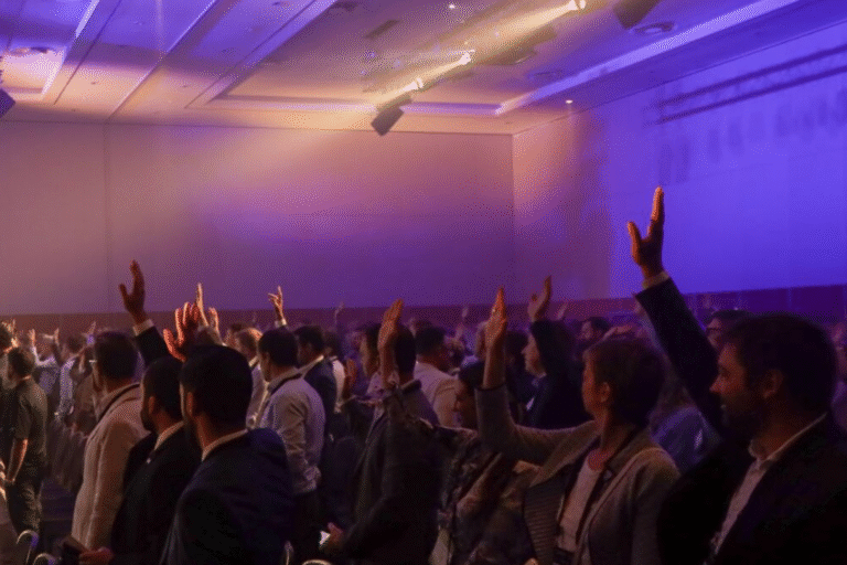Crowd of attendees with raised hands in a dimly lit indoor venue under purple stage lights.