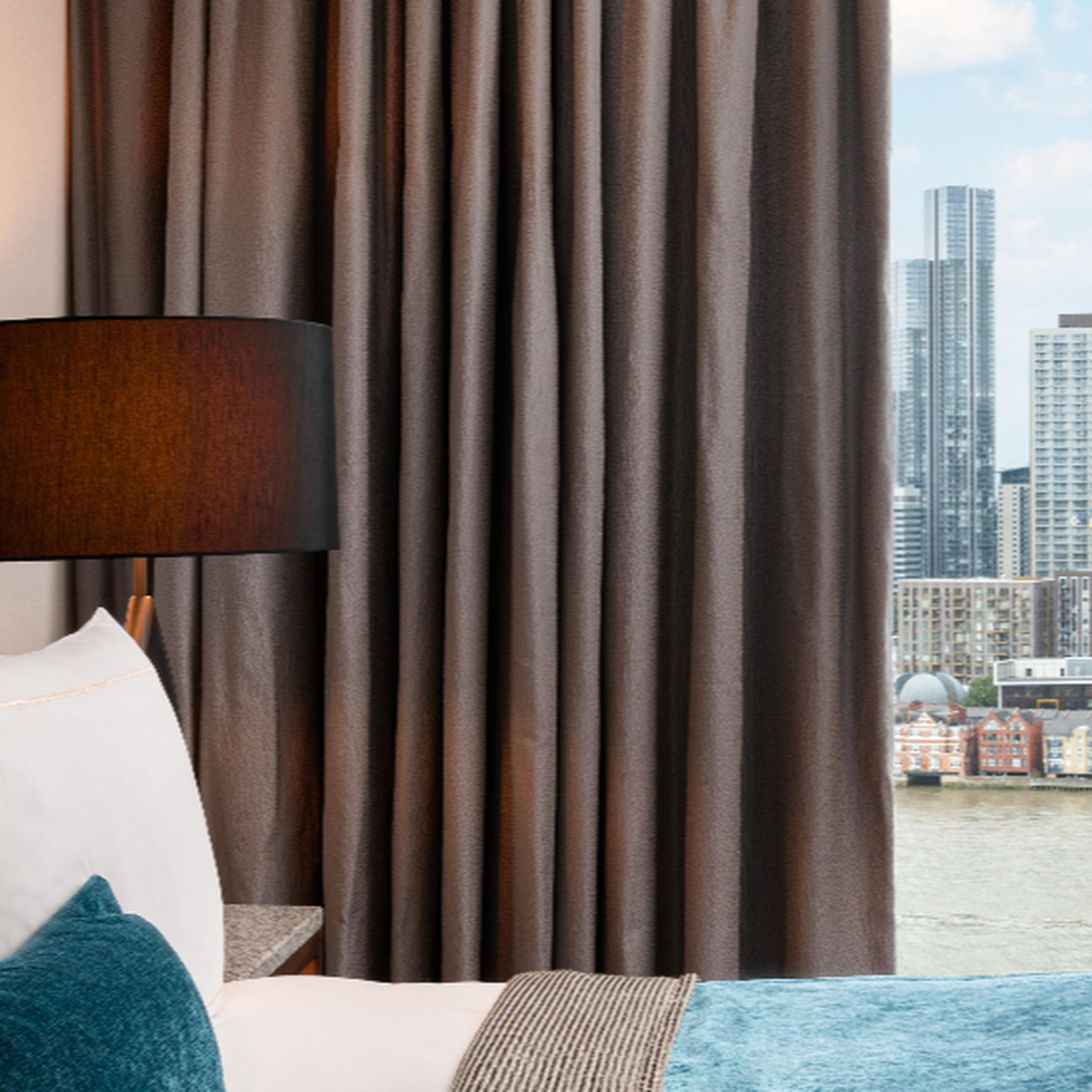 Hotel room interior with a dark lampshade, brown curtains, and a city skyline visible through the window.