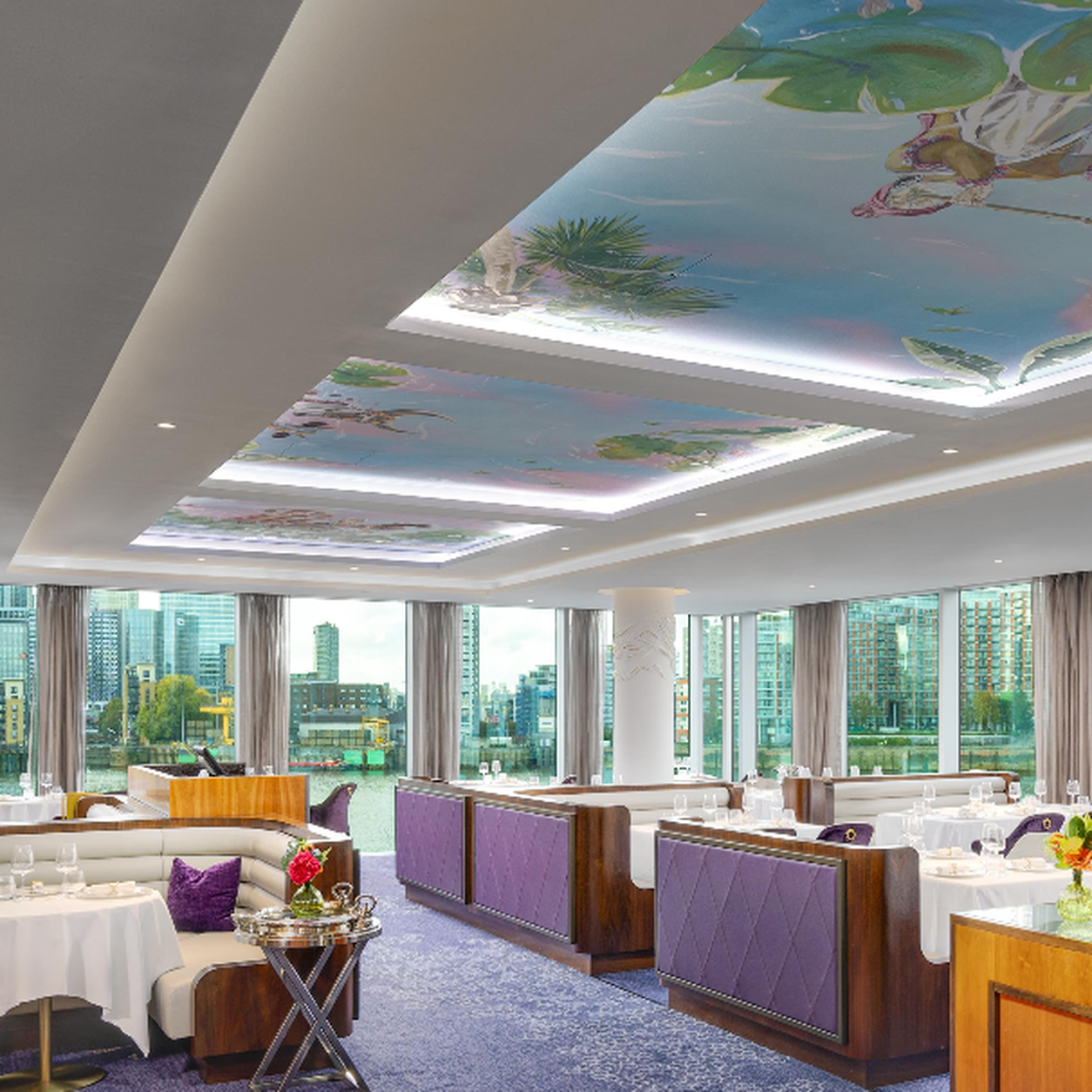 Upscale dining room with purple upholstered booths, white-clothed tables, and a tropical mural on the illuminated ceiling overlooking city windows.