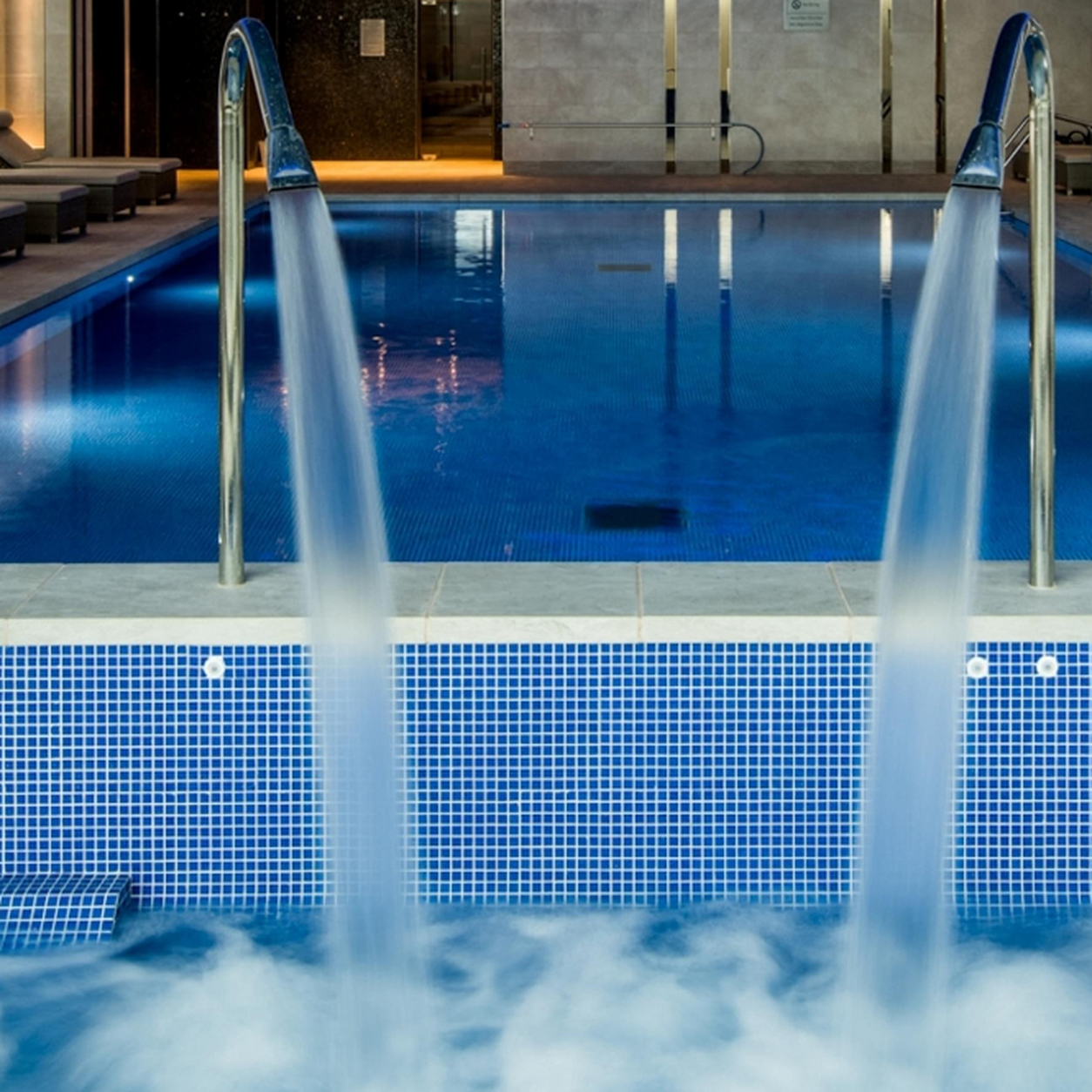 Indoor pool with two water fountain jets pouring into a blue tiled pool, lounge seating along the wall nearby.