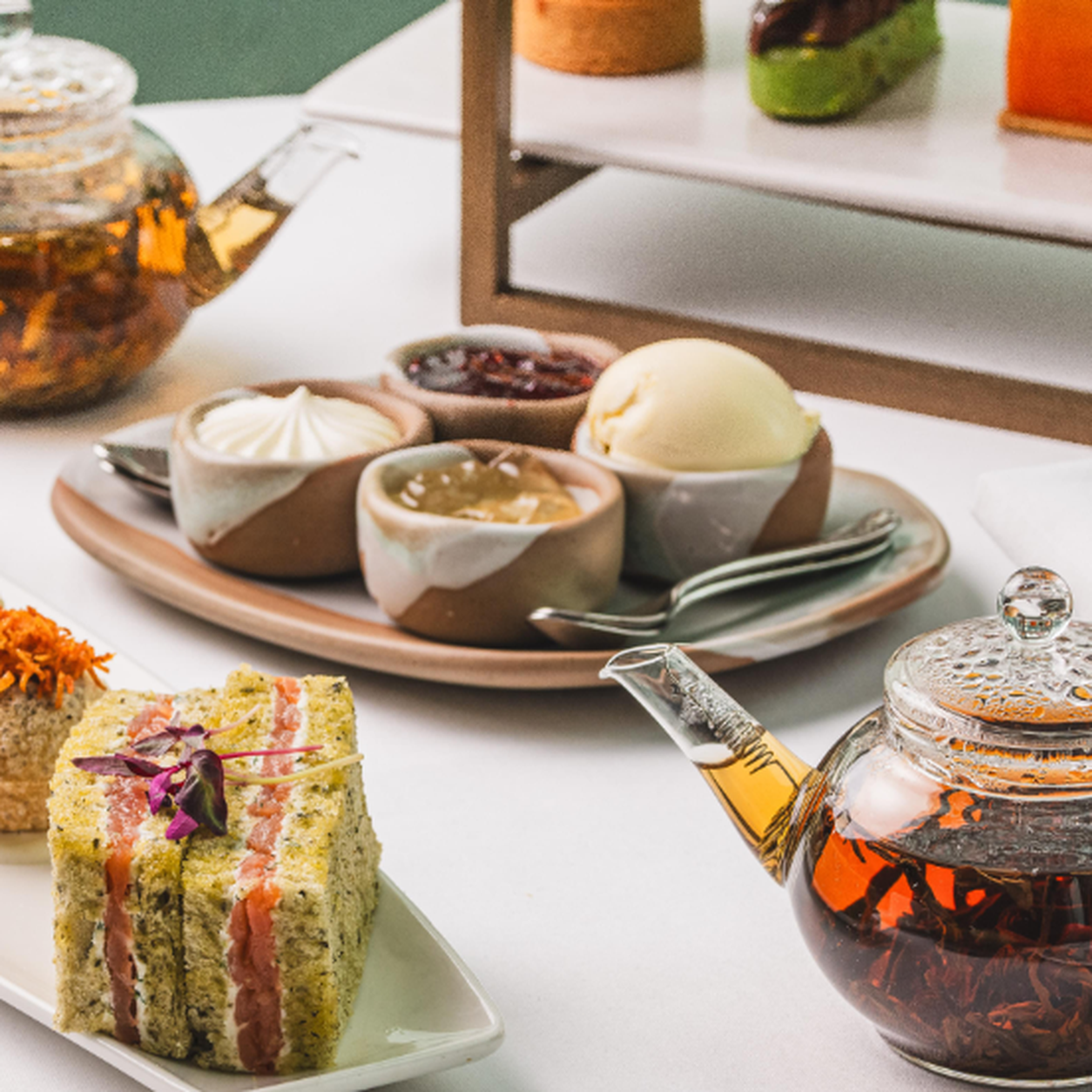 Assorted desserts in ceramic bowls on a serving plate with a glass teapot nearby on a white table.