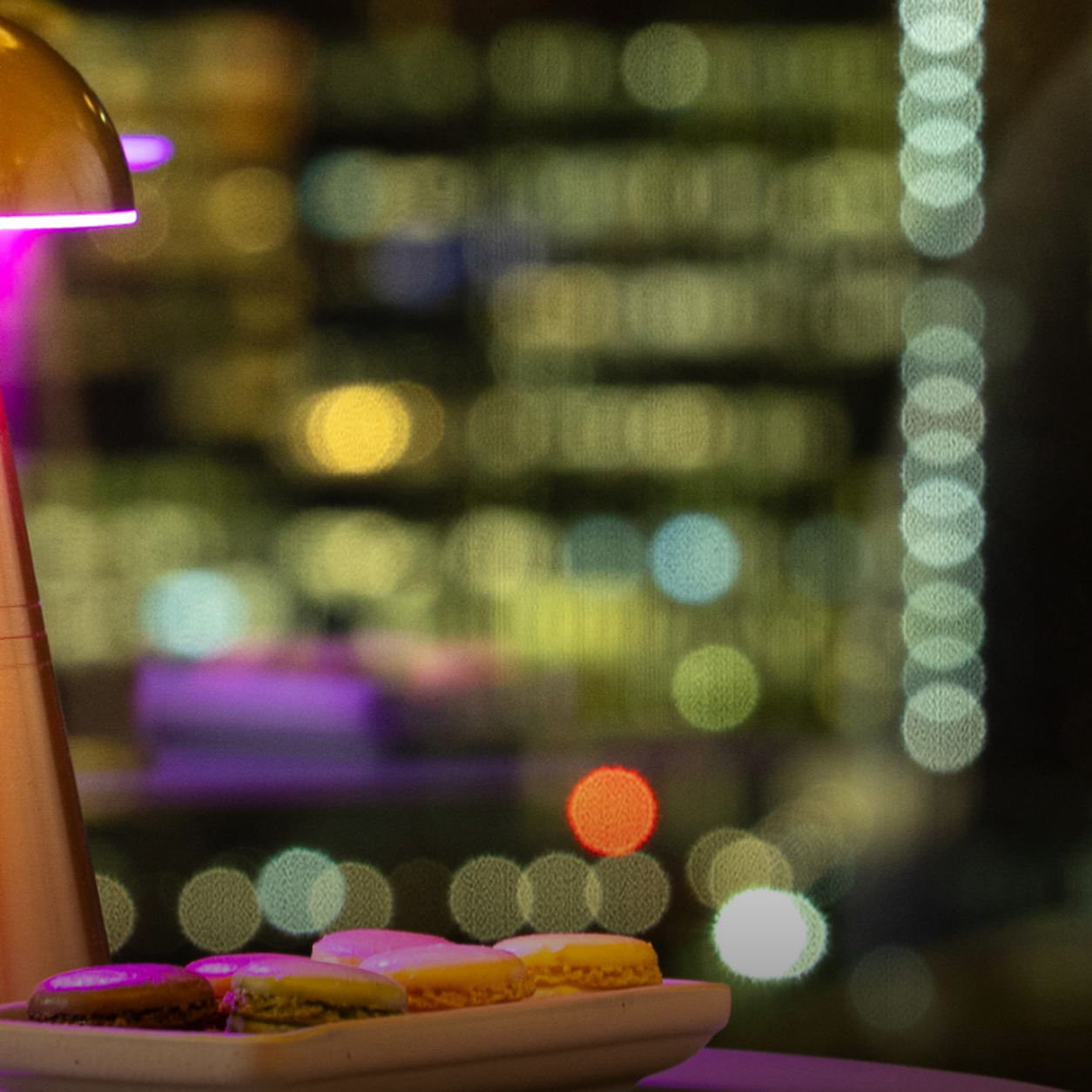 Plate of colorful macarons on a white dish with a blurred nighttime cityscape and pink lamp glow in the background
