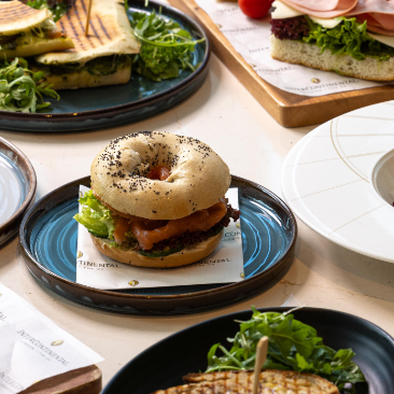 Bagel sandwich with beef patty, lettuce and sauce on a blue plate with a napkin underneath, in a casual dining spread