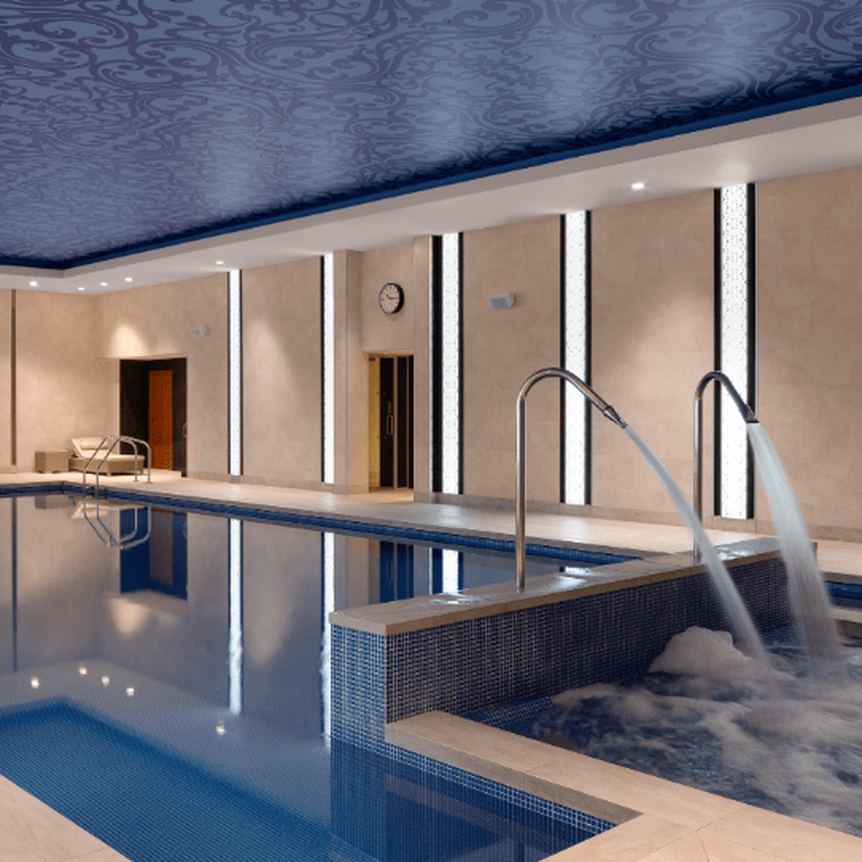 Indoor pool area with blue-tiled pool, water jets splashing into the basin, and beige walls with vertical light panels at the sides.
