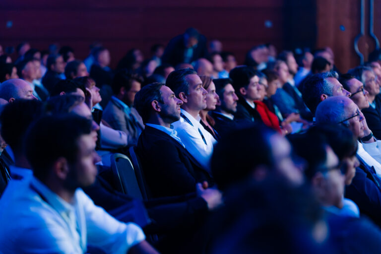 Audience seated in a dim blue-lit conference hall, focused on a presentation off-camera.