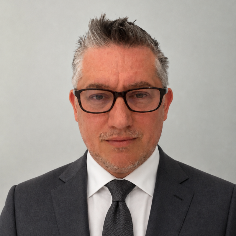 Professional head-and-shoulders portrait of a man in a dark gray suit, white shirt, and black tie, wearing glasses, facing the camera.