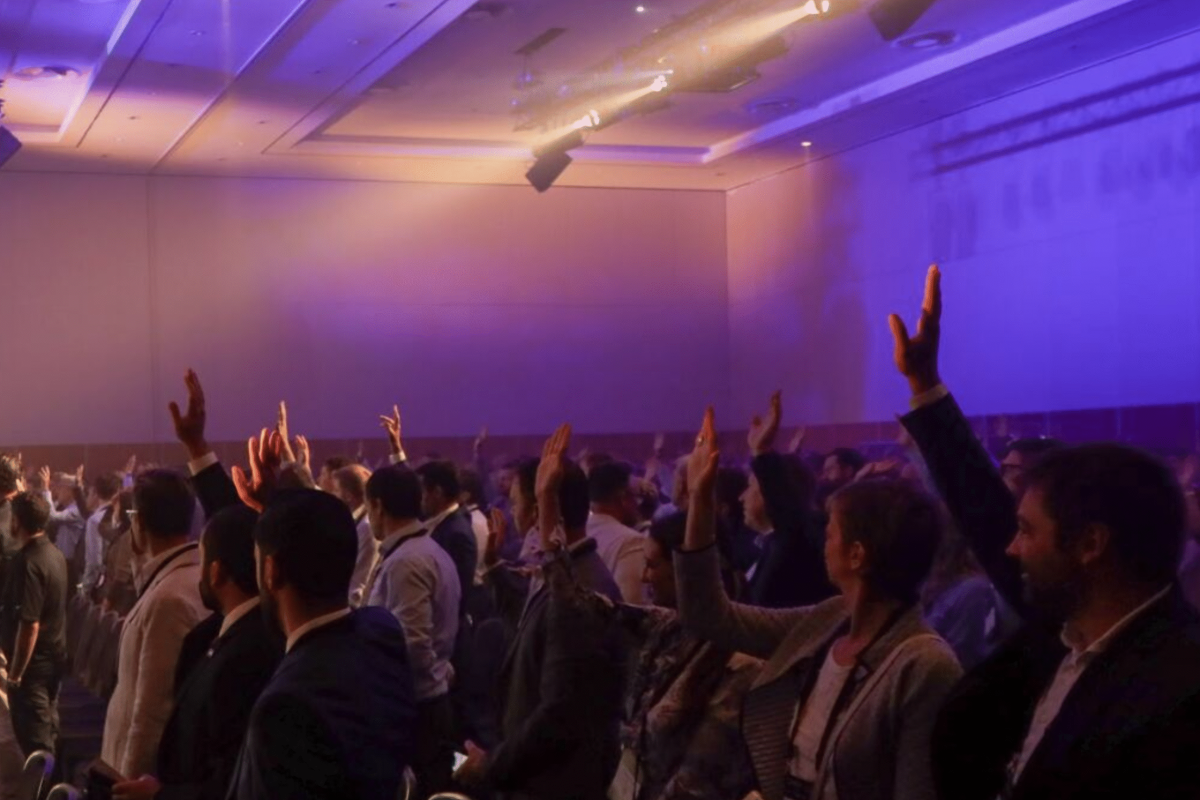 Crowd of attendees with raised hands in a dimly lit indoor venue under purple stage lights.