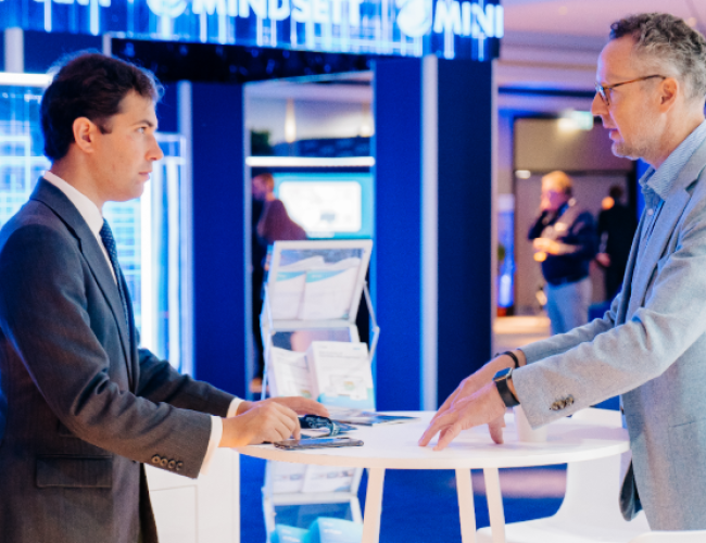 Two men in business attire discuss at a white high table at a tech trade show booth, with brochures nearby and blue lighting in the background, branding visible.